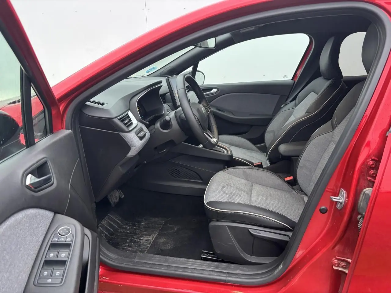 Vue intérieure côté conducteur de la Renault Clio V rouge flamme, montrant sièges gris et tableau de bord moderne.