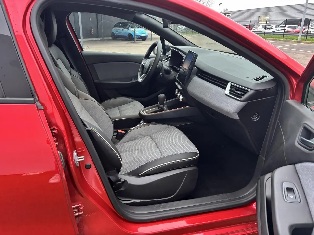 Vue intérieure côté gauche de la Renault Clio V rouge flamme, montrant sièges gris et tableau de bord avec écran tactile.