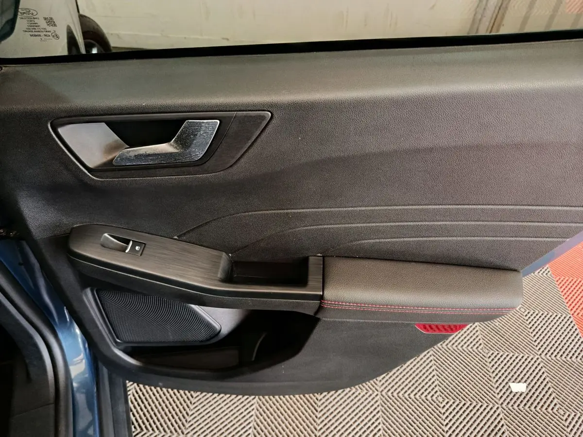 Gros plan sur la porte avant gauche du Ford Kuga bleu 2024, avec poignée argentée et surpiqûres rouges sur l'accoudoir.