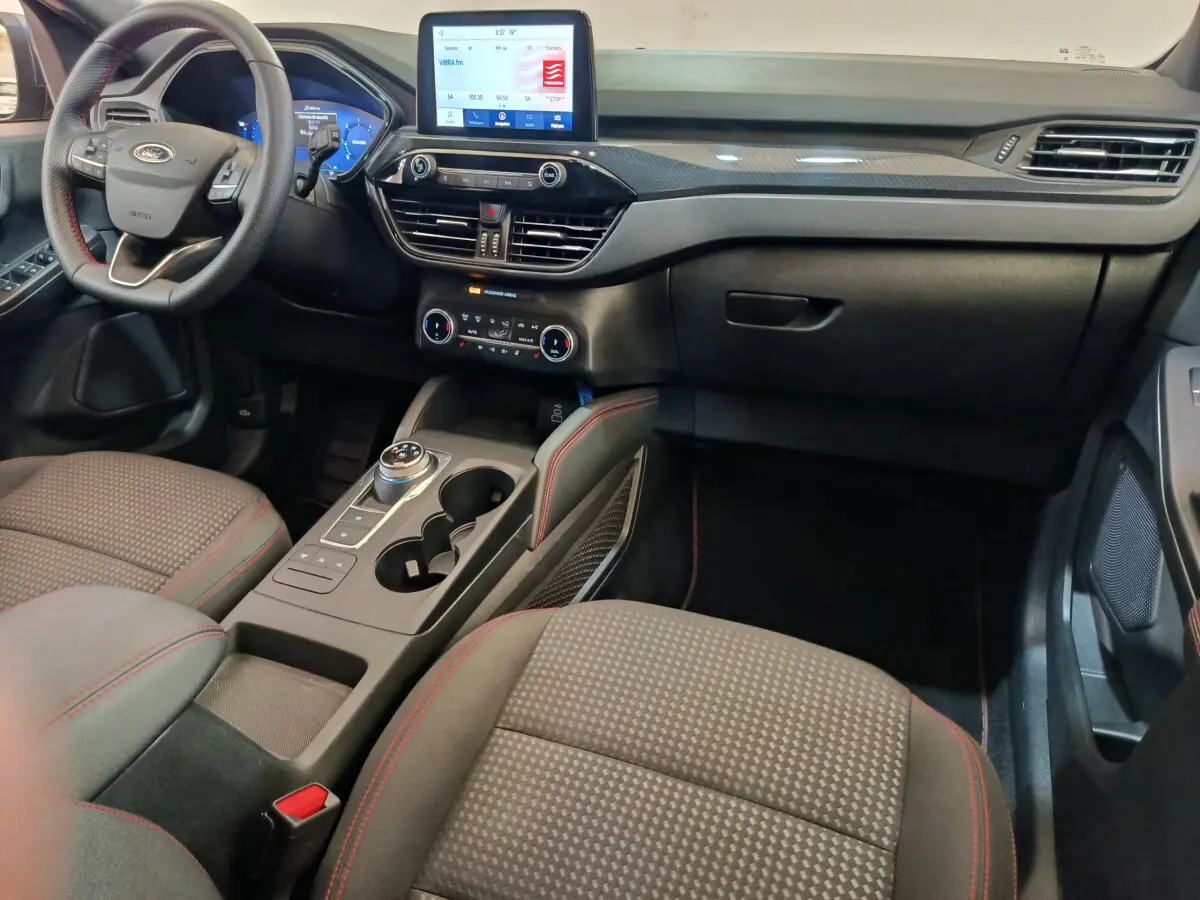 Intérieur avant du Ford Kuga 2024 St Line, tableau de bord noir avec écran tactile et sièges tissu gris à surpiqûres rouges.