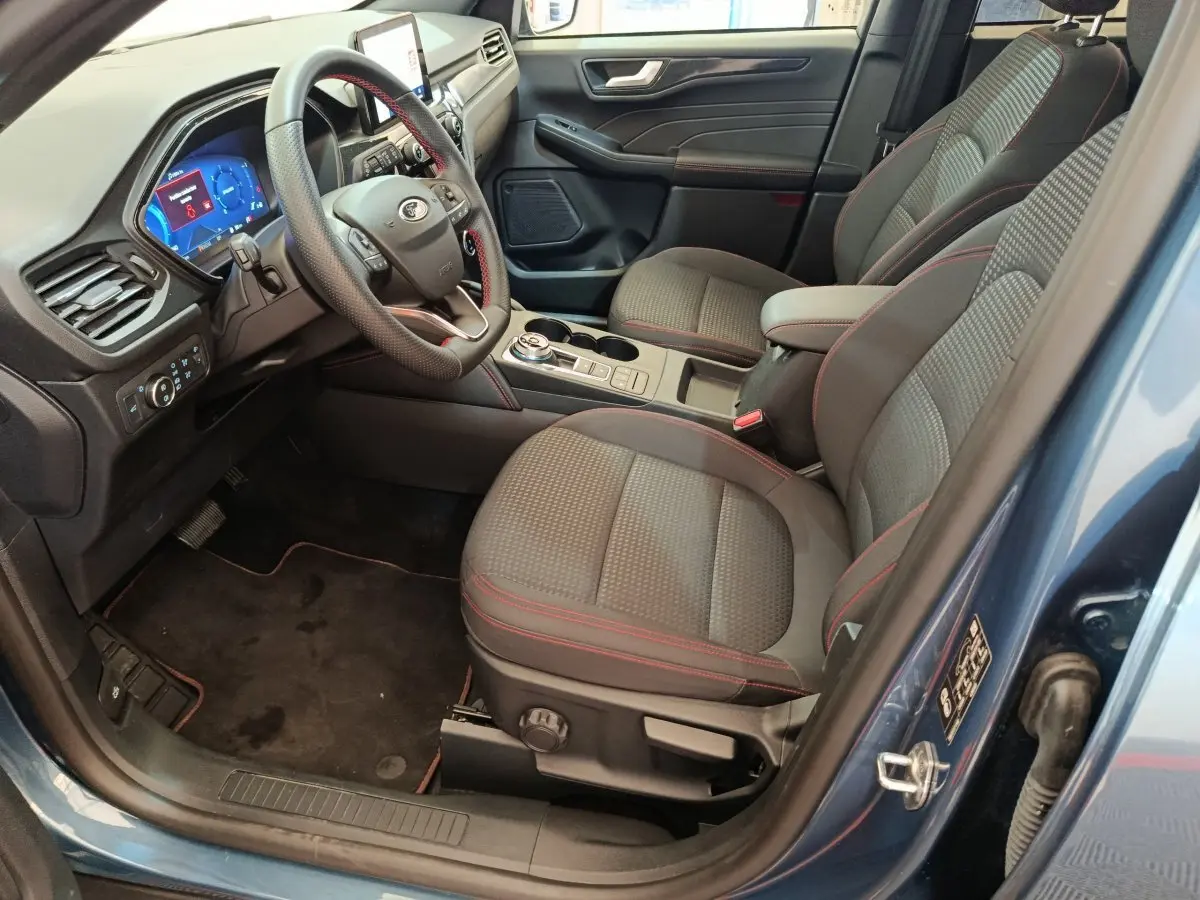 Vue intérieure côté conducteur du Ford Kuga bleu 2024, sièges tissu avec surpiqûres rouges et tableau de bord digital.