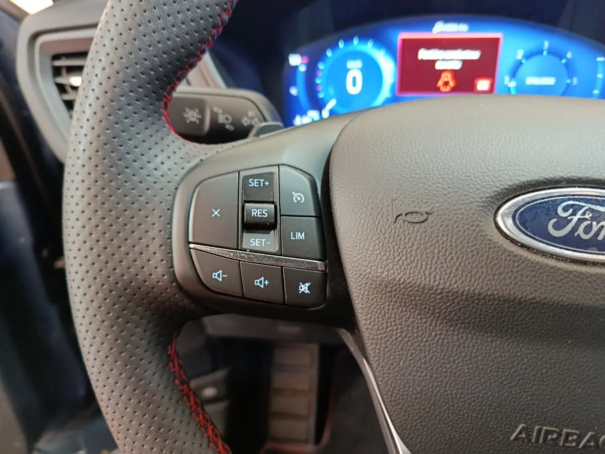 Gros plan sur le volant cuir perforé avec commandes multifonctions du Ford Kuga 2024, tableau de bord numérique bleu en arrière-plan.