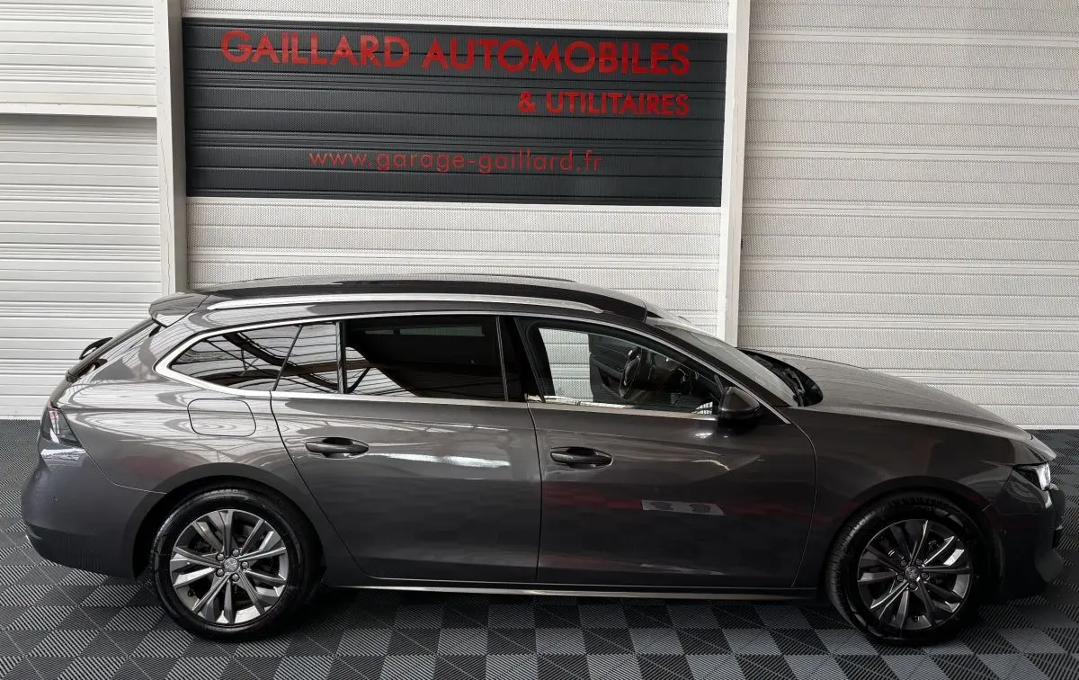 Profil droit d'une Peugeot 508 break gris foncé stationnée devant un garage avec sol damier noir et gris.