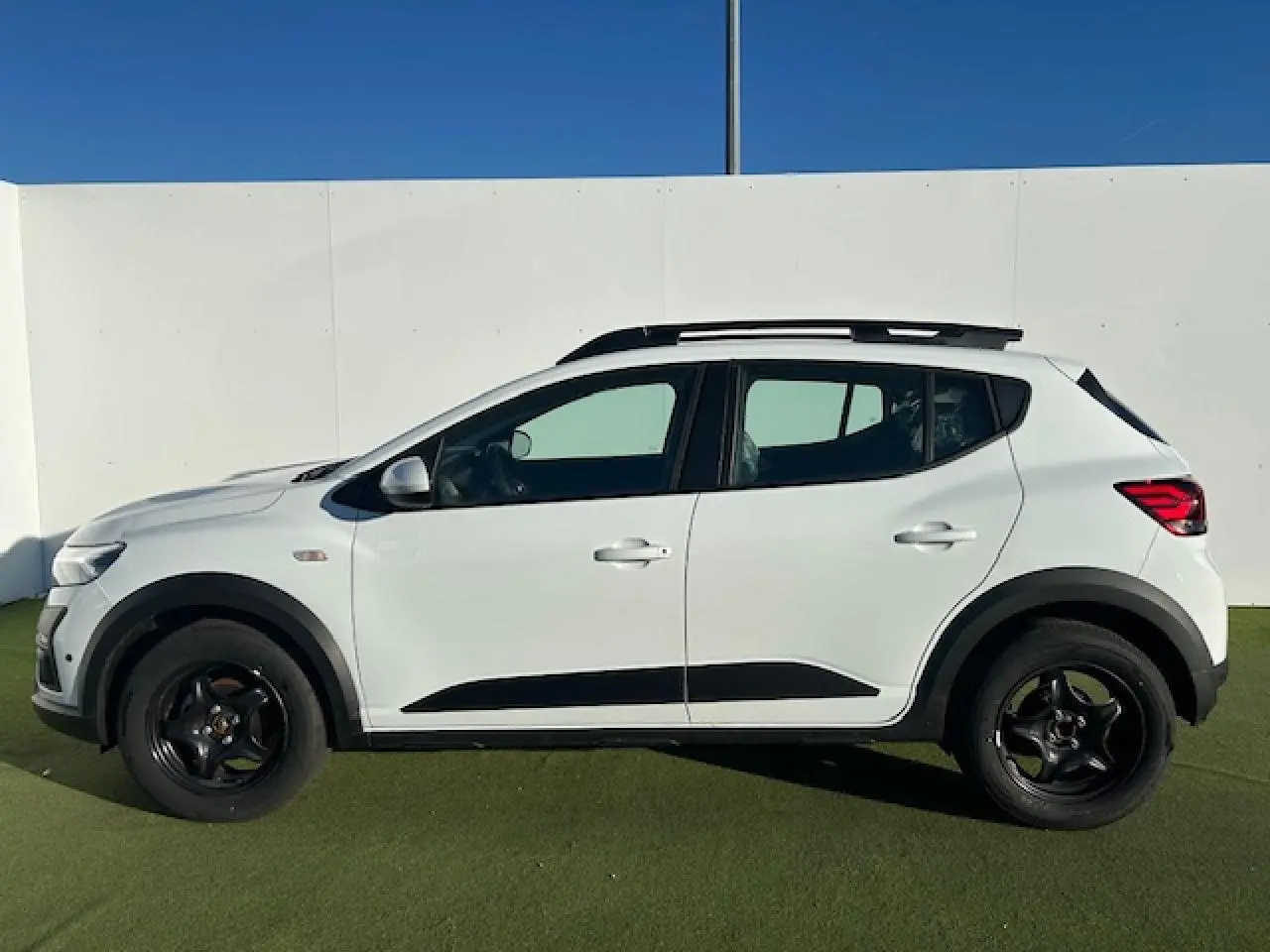 Vue latérale droite d'une Dacia Sandero Stepway blanche Arctique avec barres de toit noires et jantes foncées.