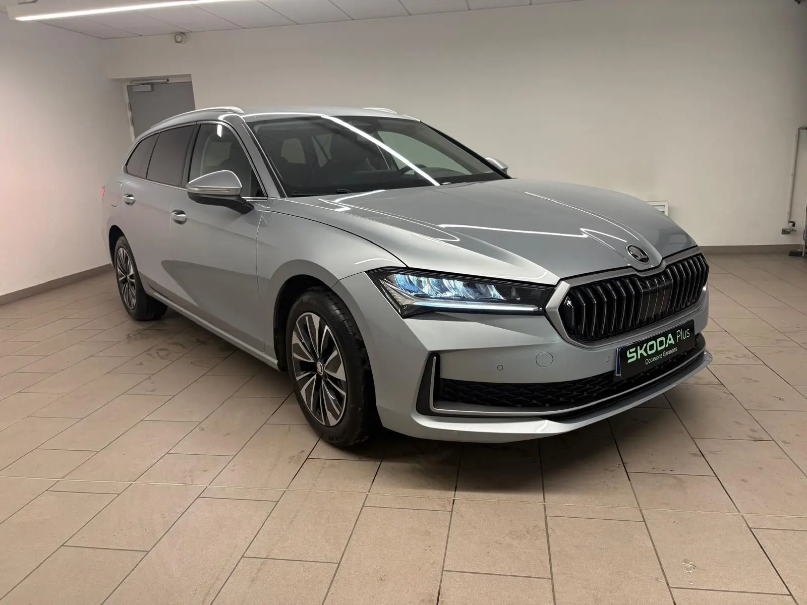 Skoda Superb Combi 2025 gris argent en 3/4 avant droit, avec calandre noire et phares LED allumés.