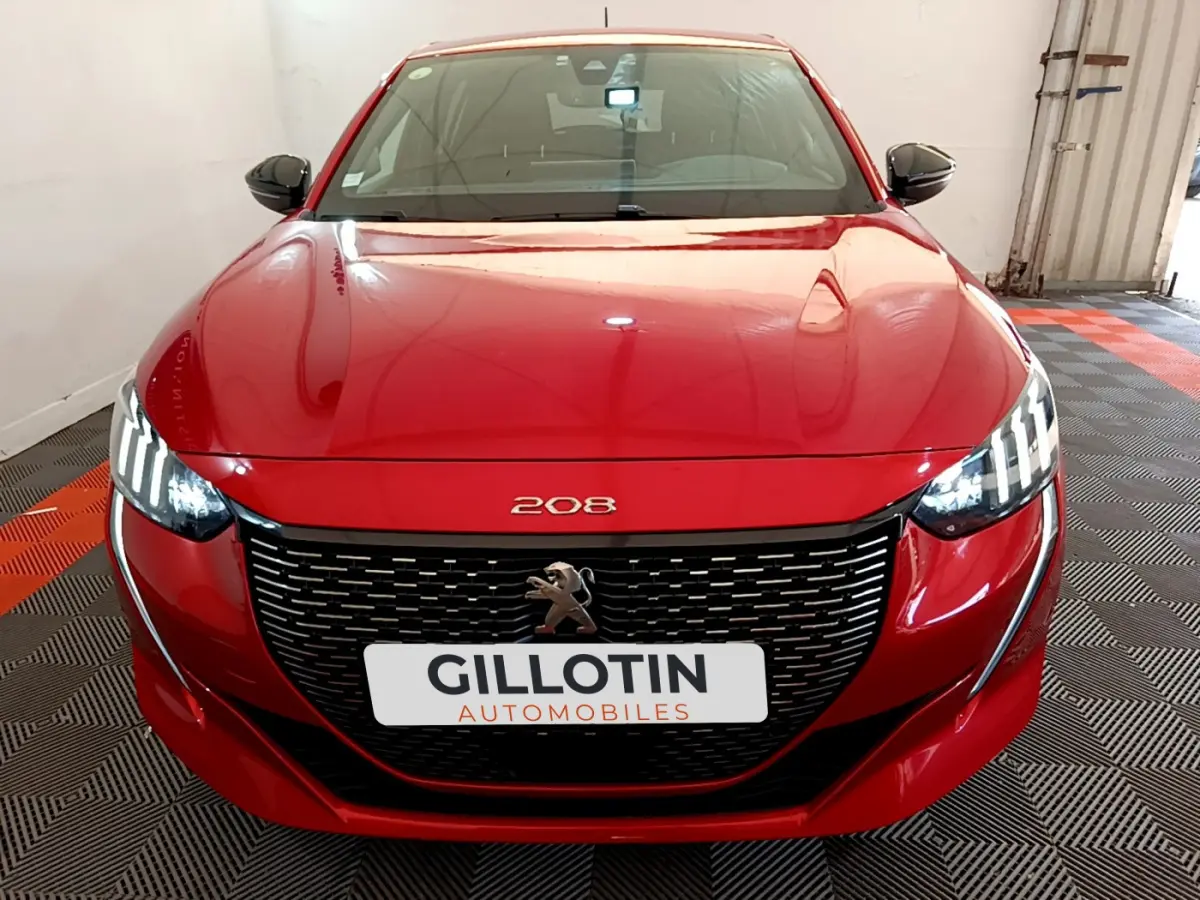 Peugeot 208 rouge vue de face, avec calandre noire et phares LED distinctifs allumés dans un garage.