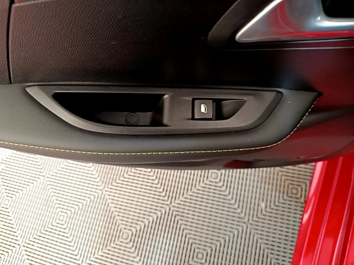 Gros plan sur la commande de lève-vitre côté gauche d'une Peugeot 208 rouge avec surpiqûres jaunes sur la portière.