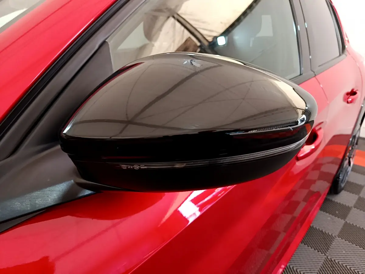 Rétroviseur noir brillant côté gauche d'une Peugeot 208 rouge GT Line vue en gros plan sur fond intérieur.