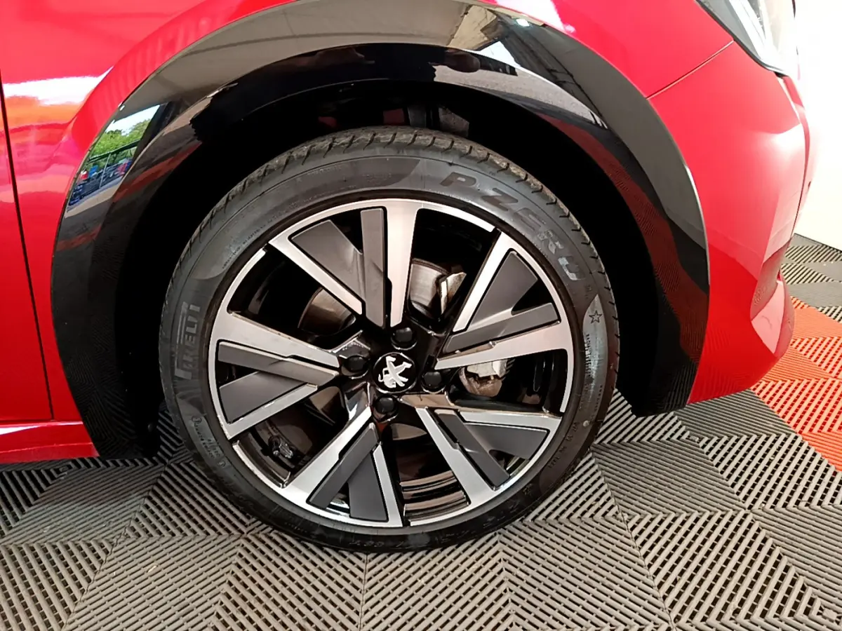 Gros plan sur la roue avant gauche d'une Peugeot 208 rouge, jante alliage noire et argentée au design moderne.