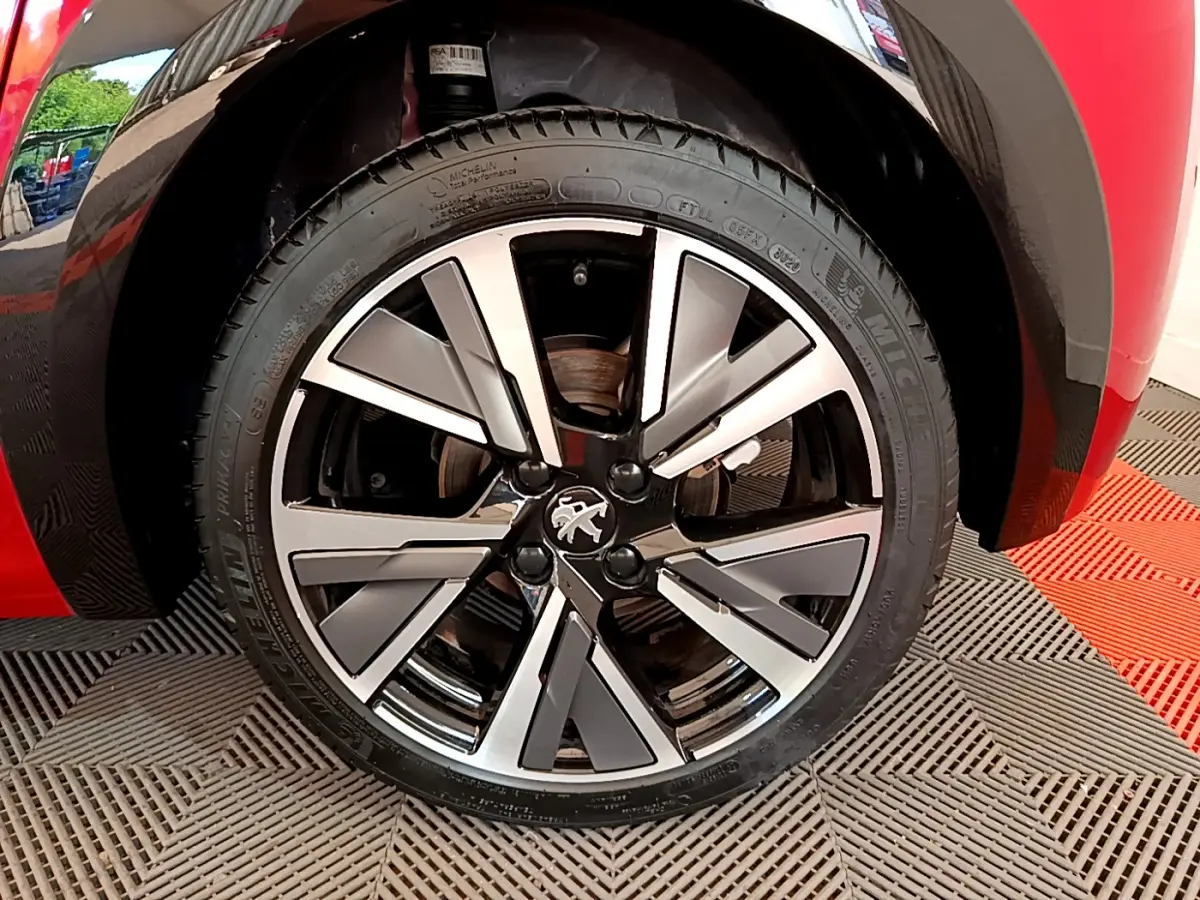Gros plan sur la roue avant gauche d'une Peugeot 208 rouge avec jante alliage au design moderne et logo lion central.