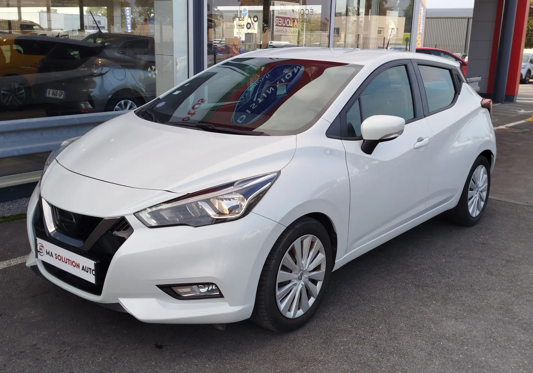 Vue 3/4 avant droit d'une Nissan Micra blanche de 2019 avec phares allumés et jantes alliage visibles.