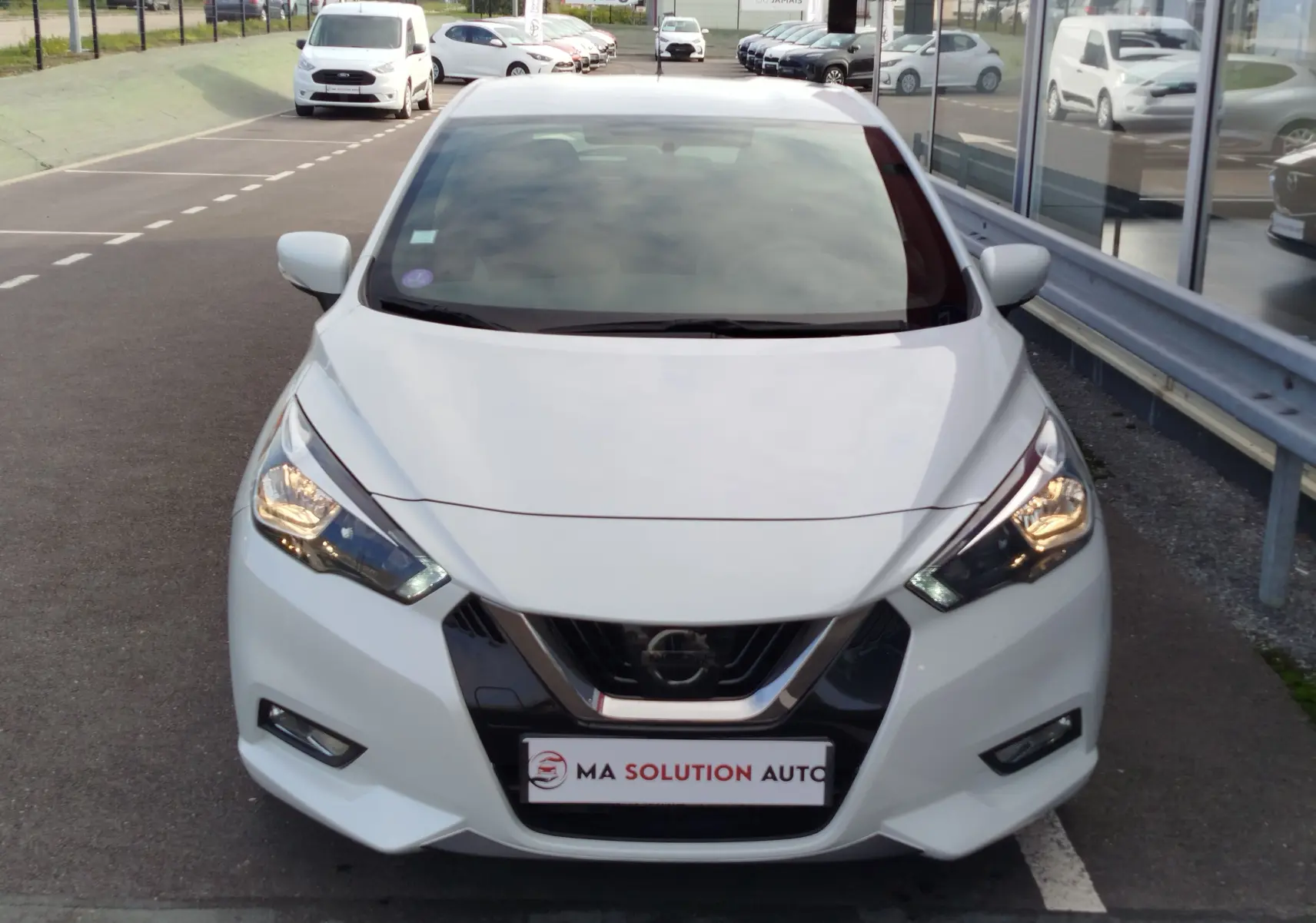 Vue frontale d'une Nissan Micra blanche 2019 avec phares allumés et calandre noire brillante.