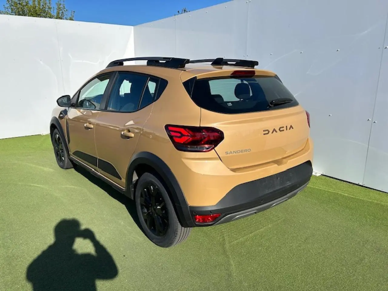 Vue 3/4 arrière droite d'une Dacia Sandero beige Safari avec barres de toit noires et jantes noires.