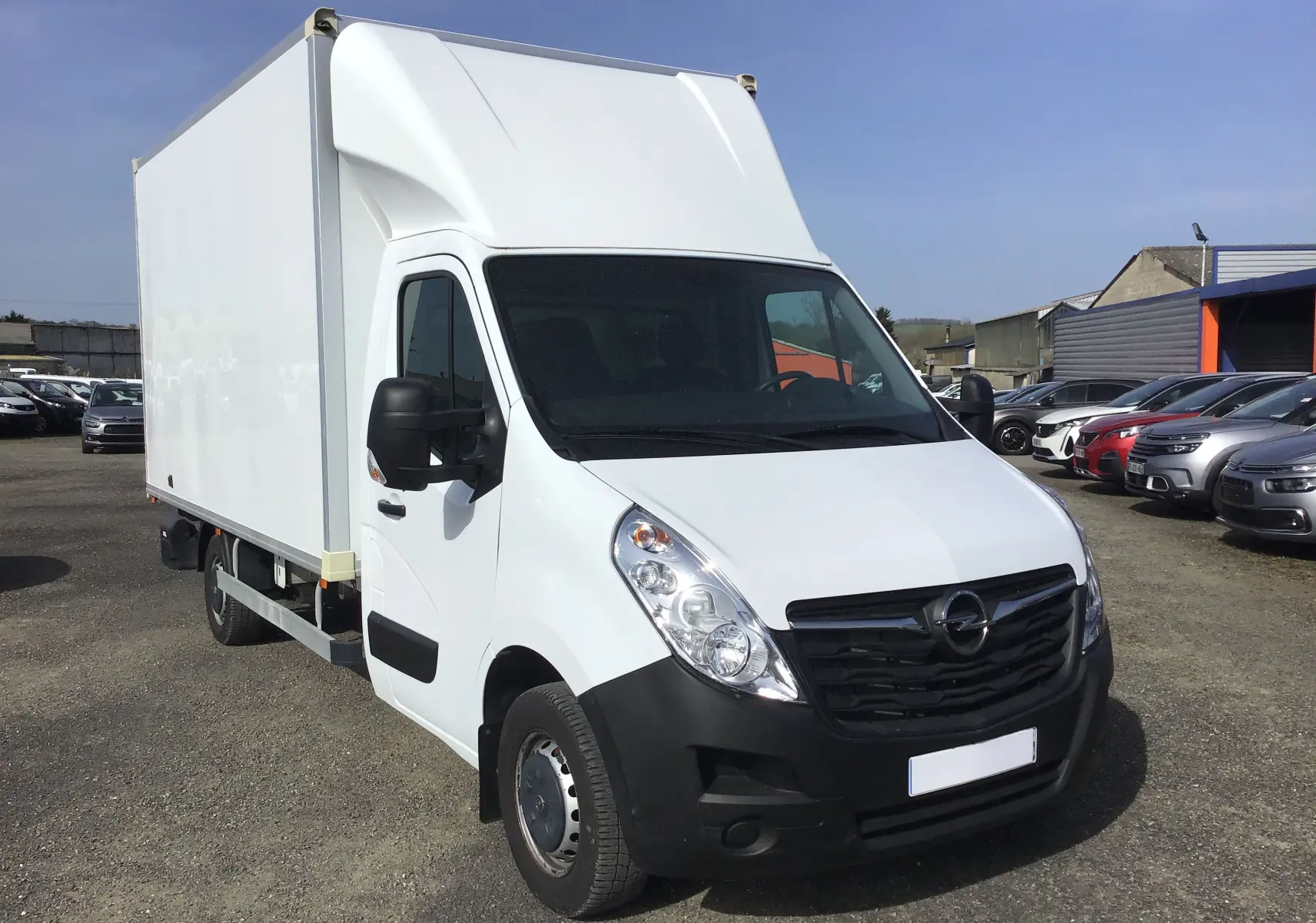 Vue 3/4 avant droite d'un Opel Movano blanc L3H1 avec caisse 20m3 et hayon, stationné en extérieur.