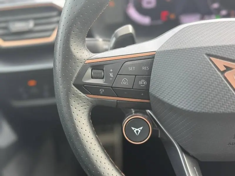 Gros plan sur le volant cuir perforé noir avec commandes multifonctions et logo CUPRA cuivré au centre.