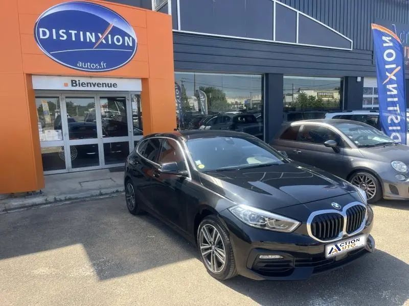 BMW Série 1 noire vue en 3/4 avant droit, avec calandre chromée et jantes alliage 16 pouces, stationnée devant un garage