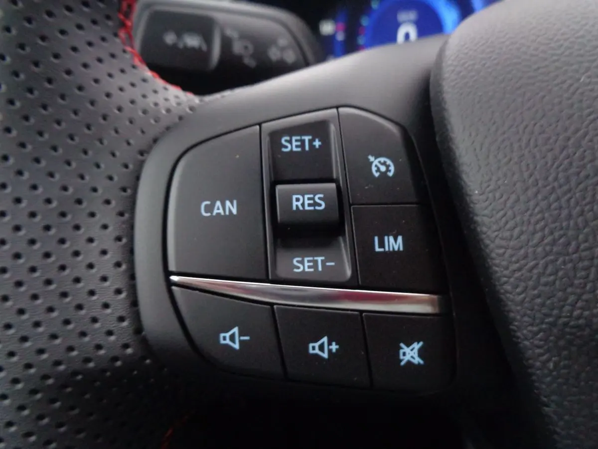 Gros plan sur les commandes du volant perforé noir du Ford Puma blanc, montrant les boutons de régulateur et volume.