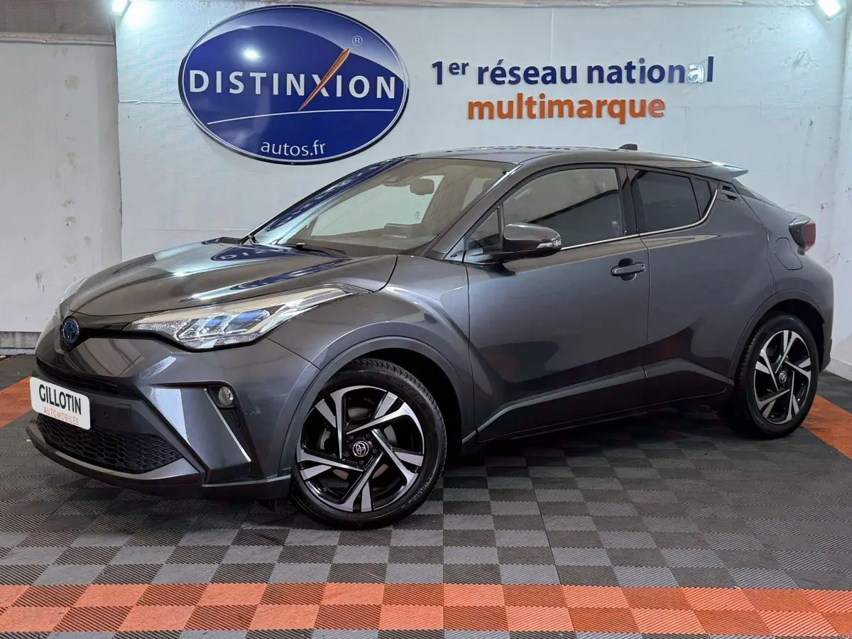 Toyota C-HR 1.8 Hybride gris foncé vu de profil avant droit avec jantes noires et logo Toyota visible.