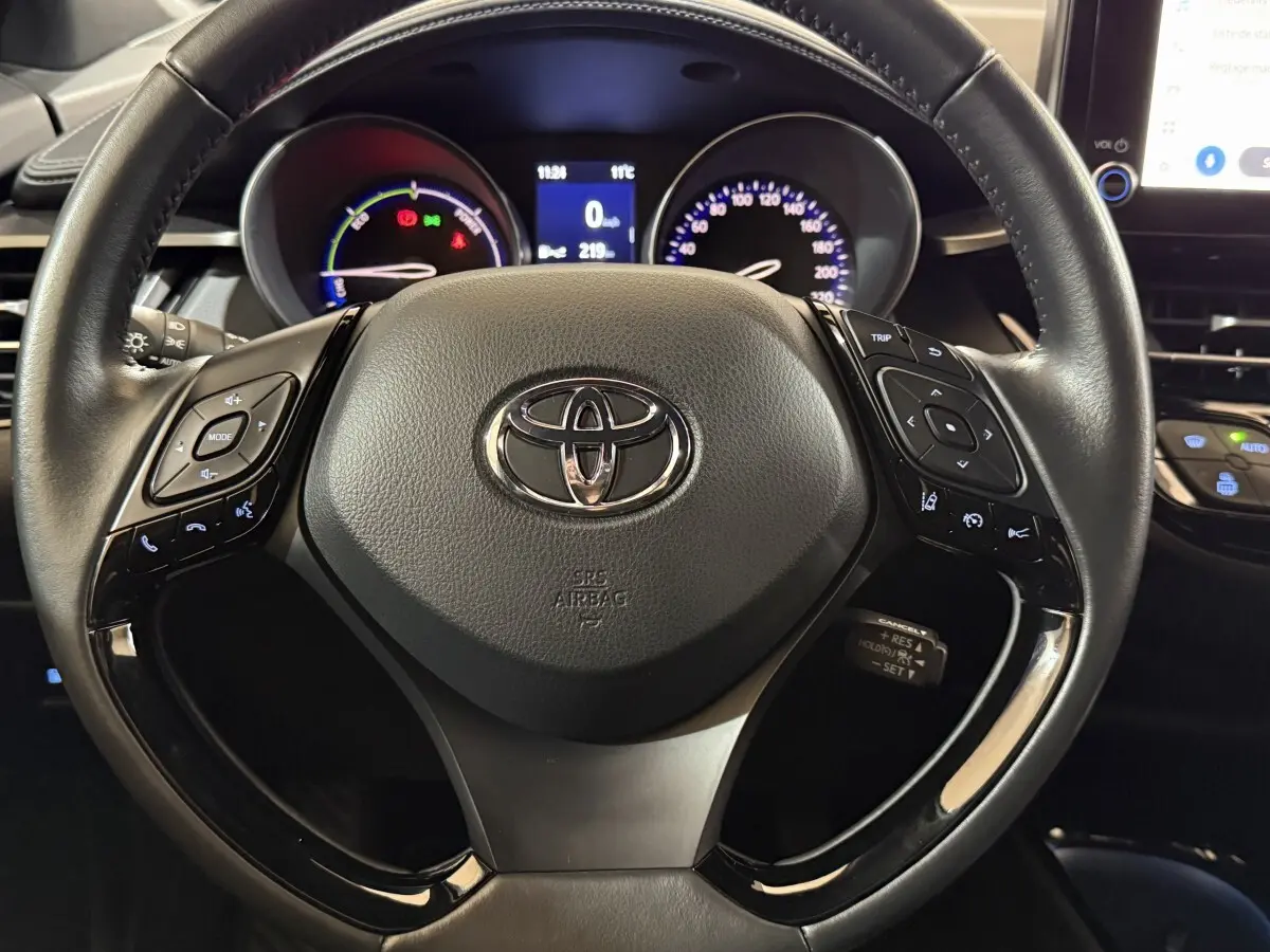 Vue rapprochée du volant cuir noir du Toyota C-HR 1.8 Hybride 122 Edition avec commandes multifonctions et tableau de bord éclairé.