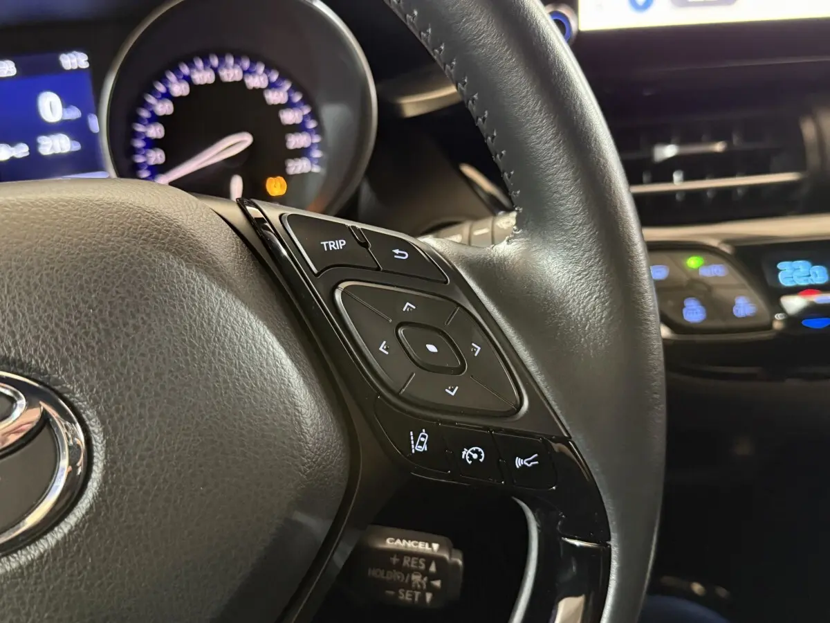 Gros plan sur le volant cuir noir du Toyota C-HR gris foncé, avec commandes multifonctions et tableau de bord lumineux en arrière-plan.