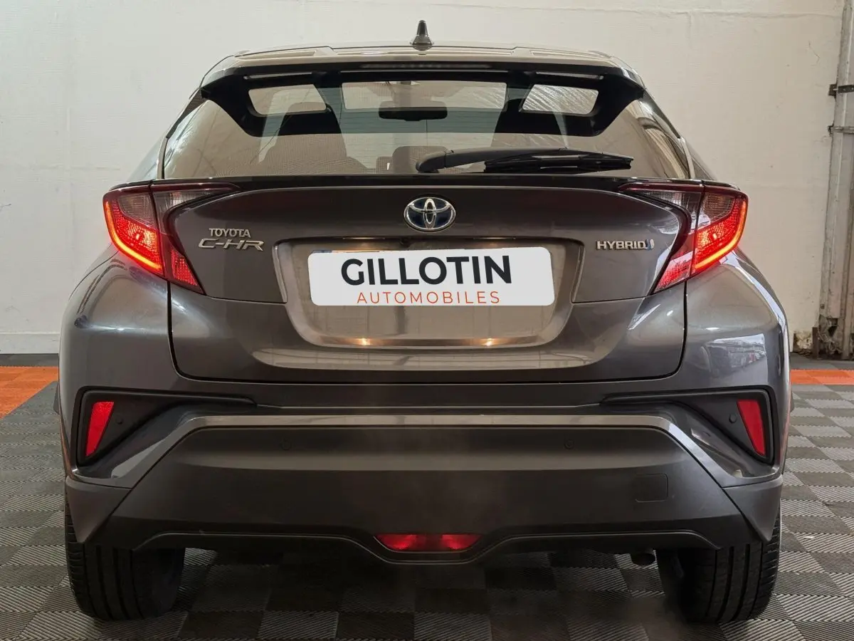 Vue arrière d'une Toyota C-HR 1.8 Hybride gris foncé avec feux arrière allumés et logo Hybrid visible.