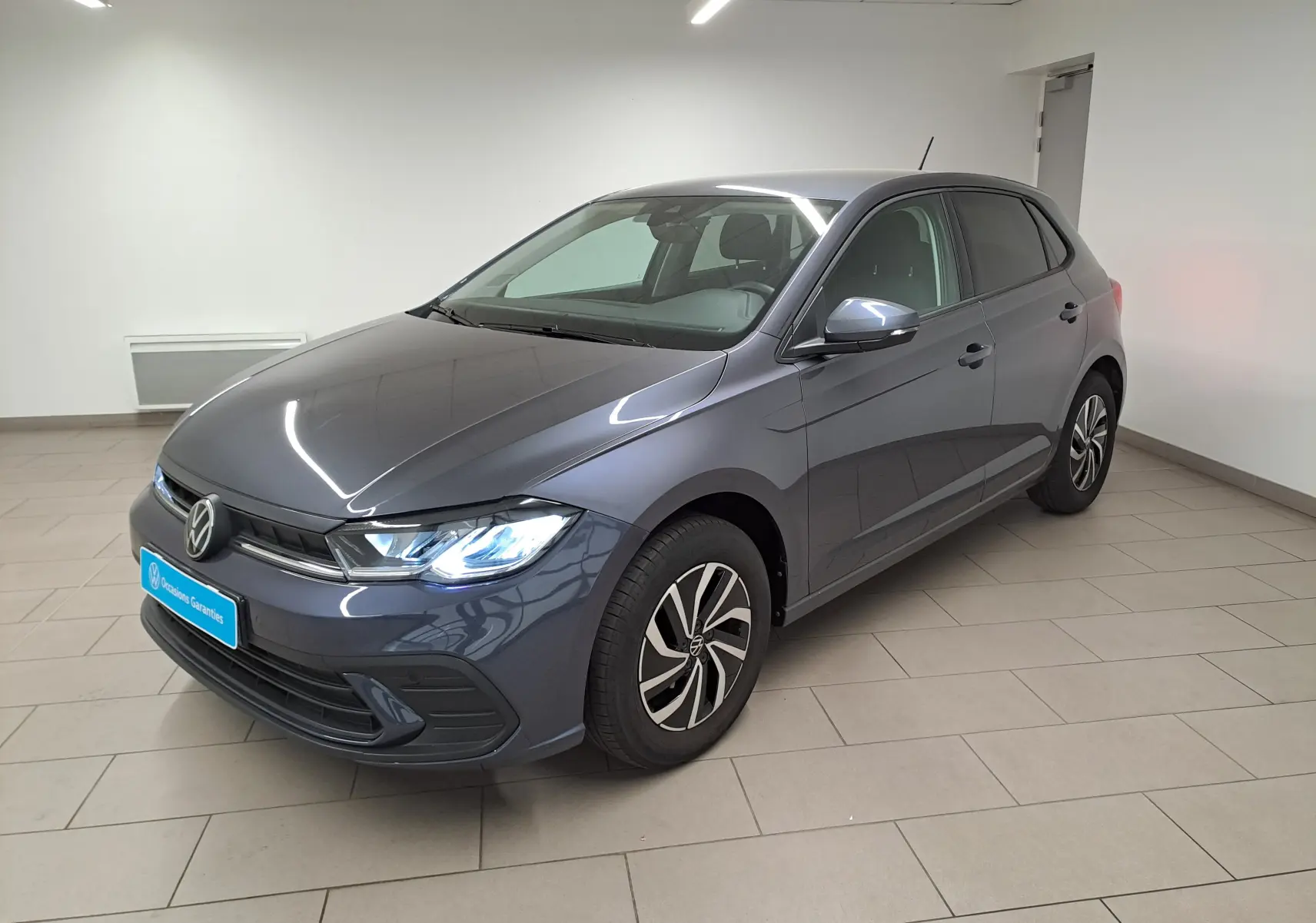Volkswagen Polo gris cendre vue 3/4 avant droit en intérieur, phares allumés et jantes spécifiques VW Edition.