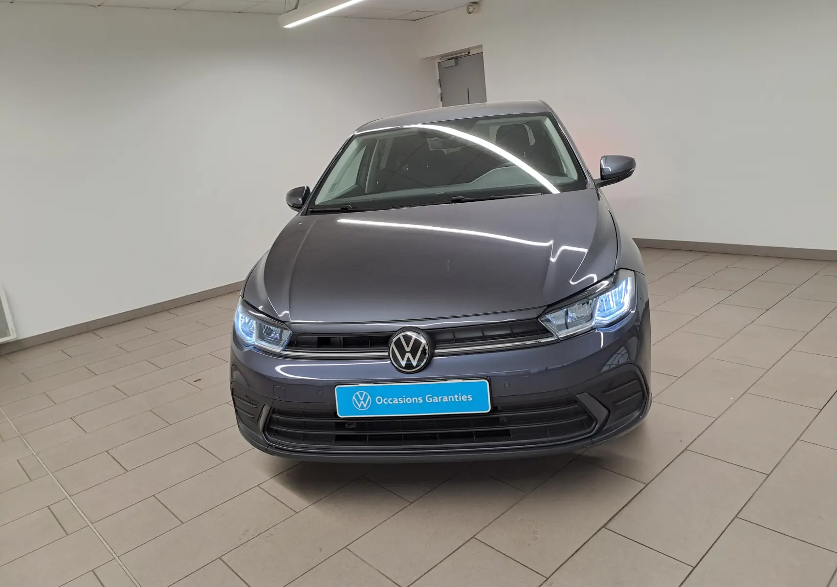 Volkswagen Polo gris cendre vue de face en intérieur avec phares allumés et plaque "Occasions Garantis" visible.