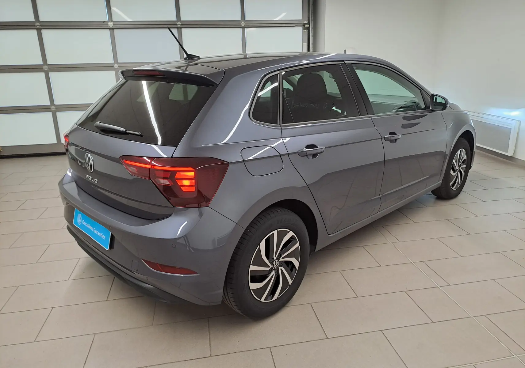 Volkswagen Polo gris cendre vue 3/4 arrière droit en intérieur, feux arrière allumés et jantes spécifiques VW Edition.