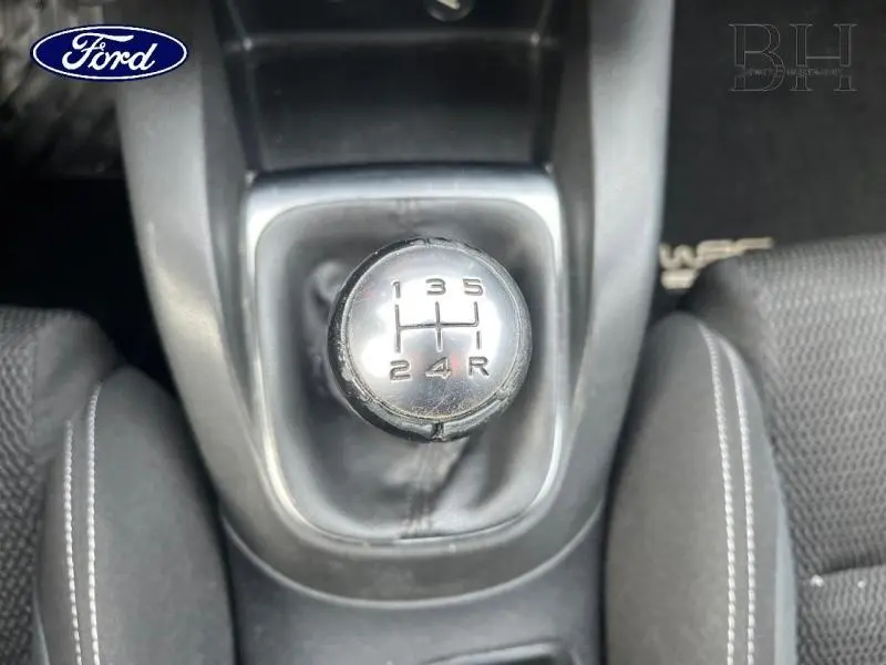 Gros plan sur le levier de vitesses manuel noir au centre de l'habitacle, entouré de sièges en tissu gris.