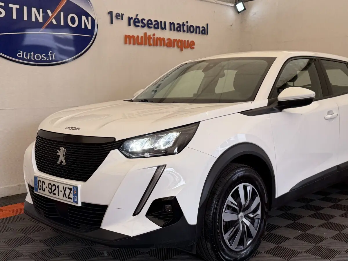 Peugeot 2008 blanc vue 3/4 avant droit, avec calandre noire et feux LED allumés en intérieur showroom.