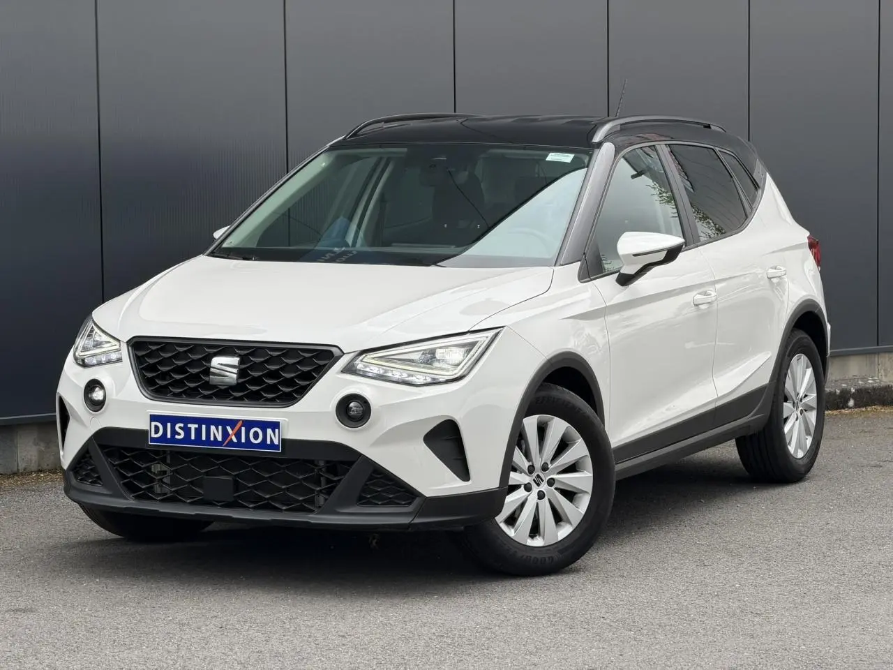 Vue 3/4 avant droite d'un SEAT Arona blanc avec toit noir et jantes alliage, stationné devant un mur gris.