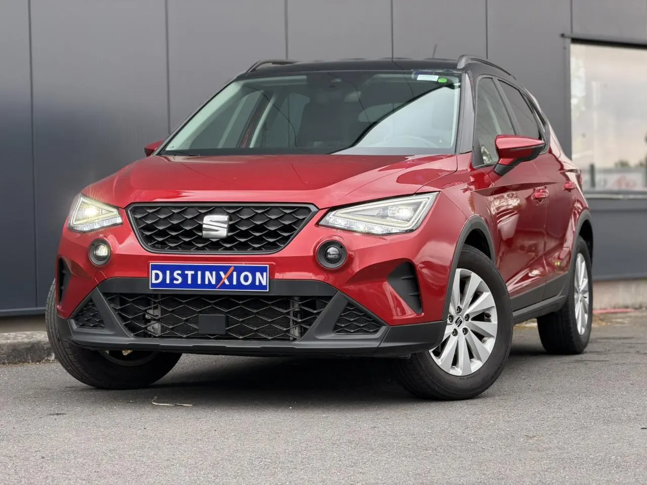 Vue 3/4 avant d'un SEAT Arona rouge Désir avec toit noir et jantes alliage, stationné devant un mur gris.