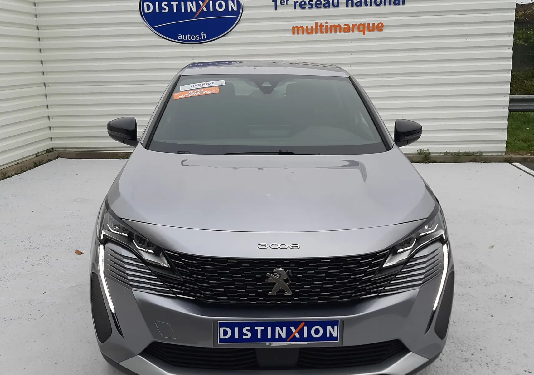 Vue frontale d'un Peugeot 3008 Hybrid gris Artense avec calandre noire et feux LED distinctifs, stationné devant un mur blanc.