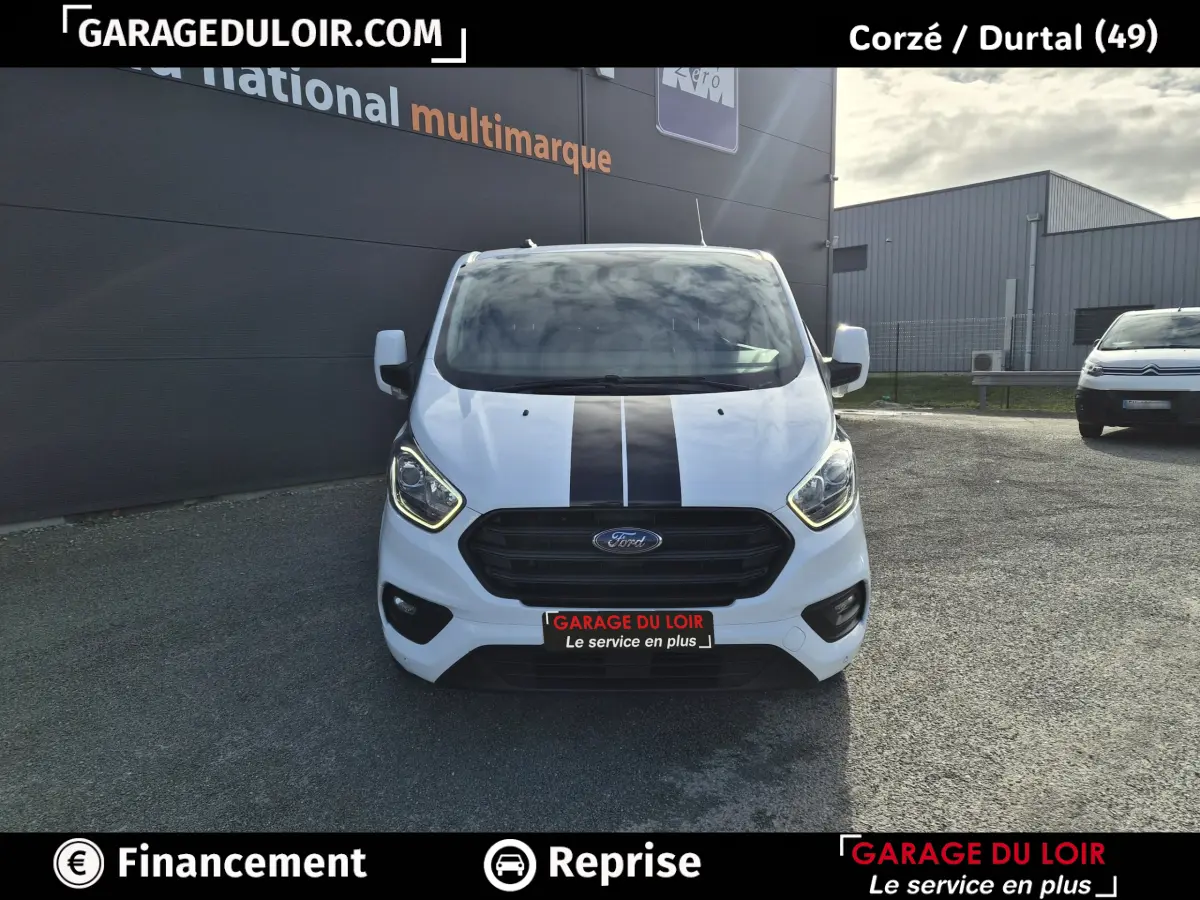 Vue avant d'un Ford Transit Custom blanc 2023 avec bande noire centrale et calandre noire, stationné devant un bâtiment.