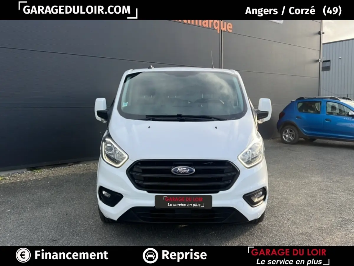Vue de face d'un Ford Transit Custom Fourgon blanc 2023 avec calandre noire et feux allumés sur parking extérieur.