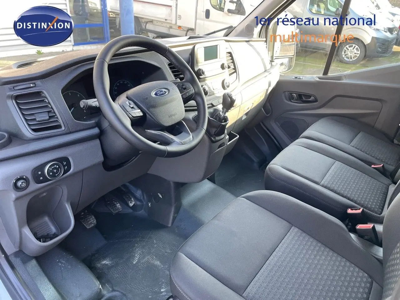 Vue intérieure côté conducteur du Ford Transit Fourgon 2023, montrant le volant, la boîte manuelle et les sièges gris.