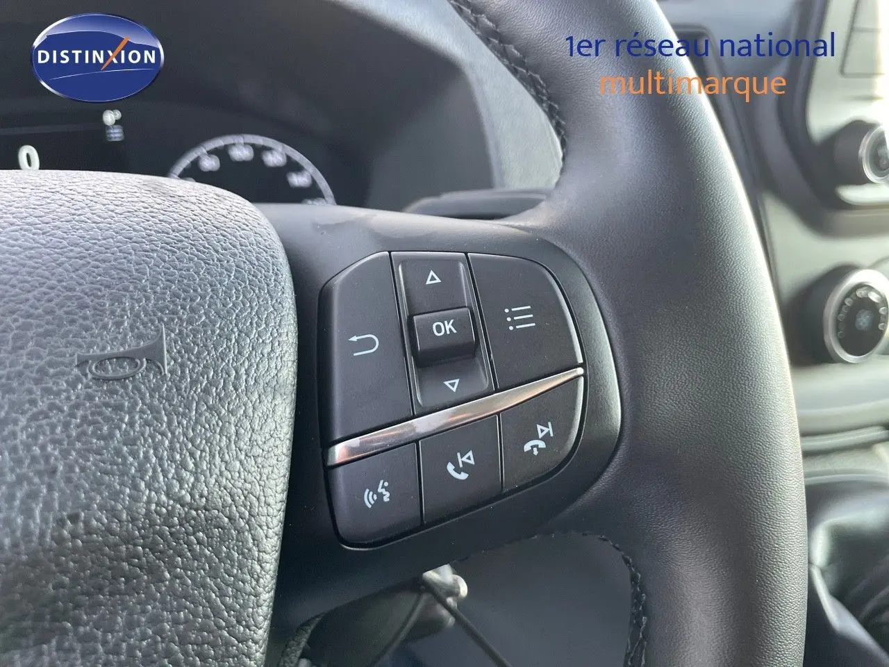 Gros plan sur les commandes multifonctions au volant noir du Ford Transit Fourgon blanc, tableau de bord en arrière-plan.