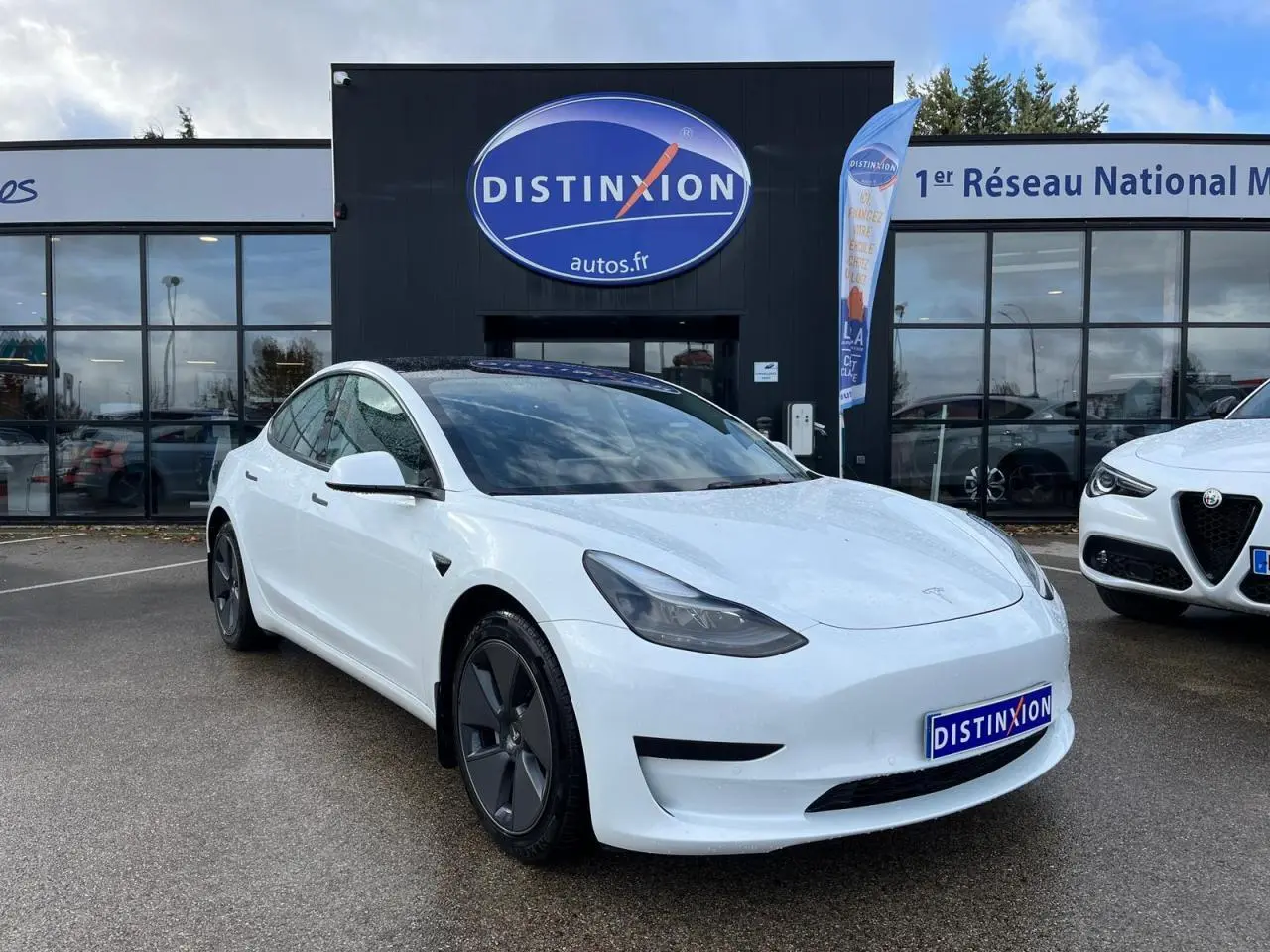 Tesla Model 3 blanc en 3/4 avant droit, avec toit panoramique en verre et jantes aérodynamiques noires.