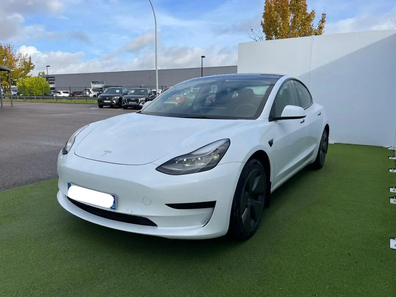 Tesla Model 3 blanc vue 3/4 avant droit avec toit panoramique en verre et jantes noires sur parking.