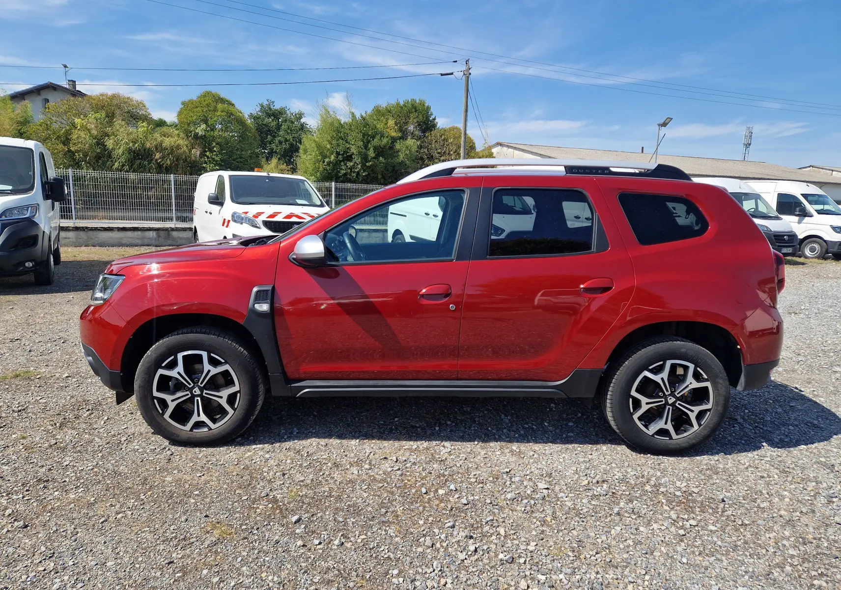 Profil droit d’un Dacia Duster 2021 rouge fusion avec jantes alliage et barres de toit visibles en extérieur.