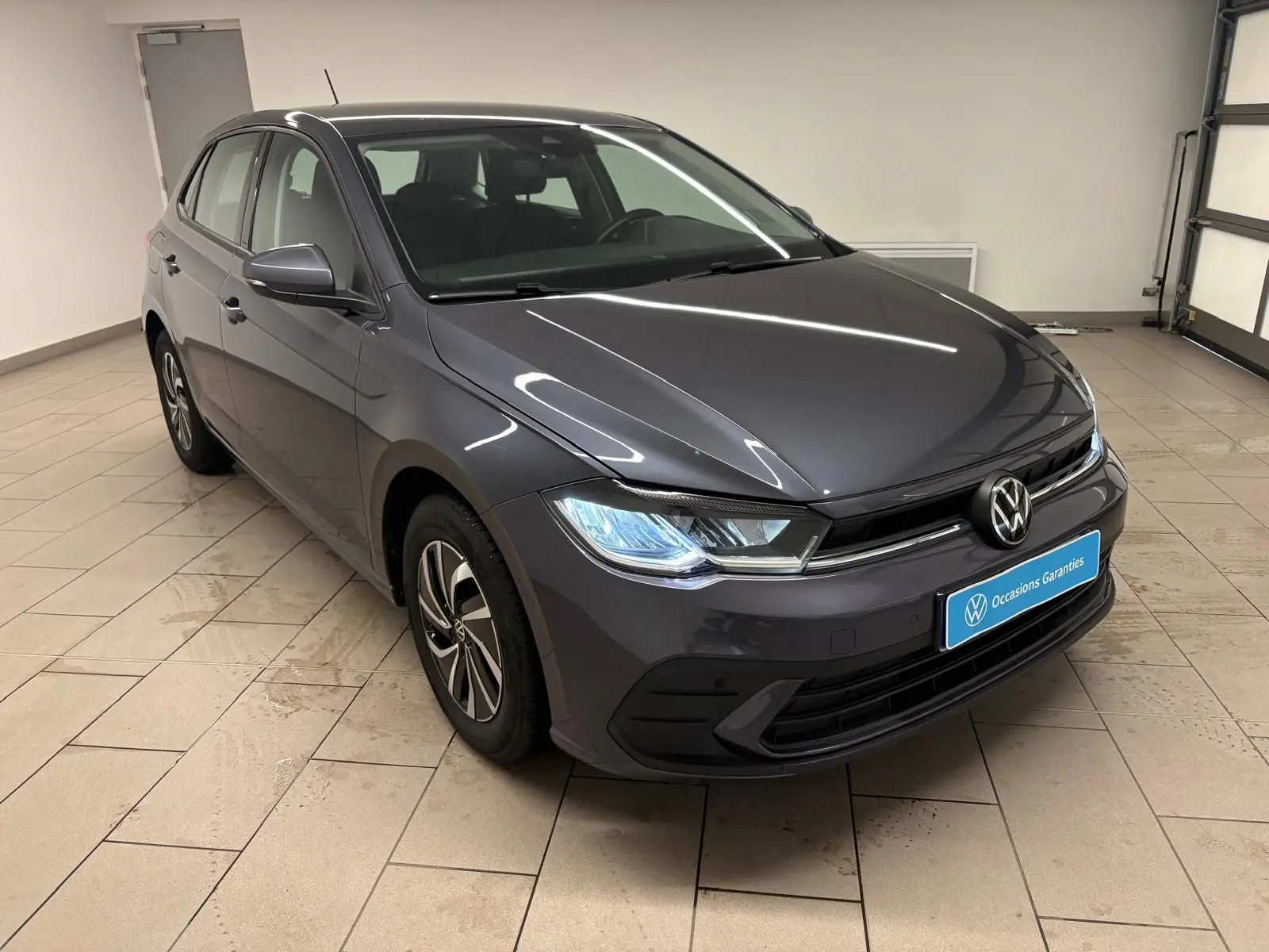 Volkswagen Polo gris cendre 2024 vue 3/4 avant droit dans un showroom avec phares allumés.