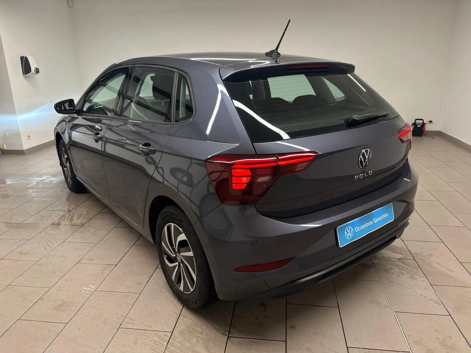 Volkswagen Polo gris cendre vue 3/4 arrière droit dans un showroom avec feux arrière allumés.