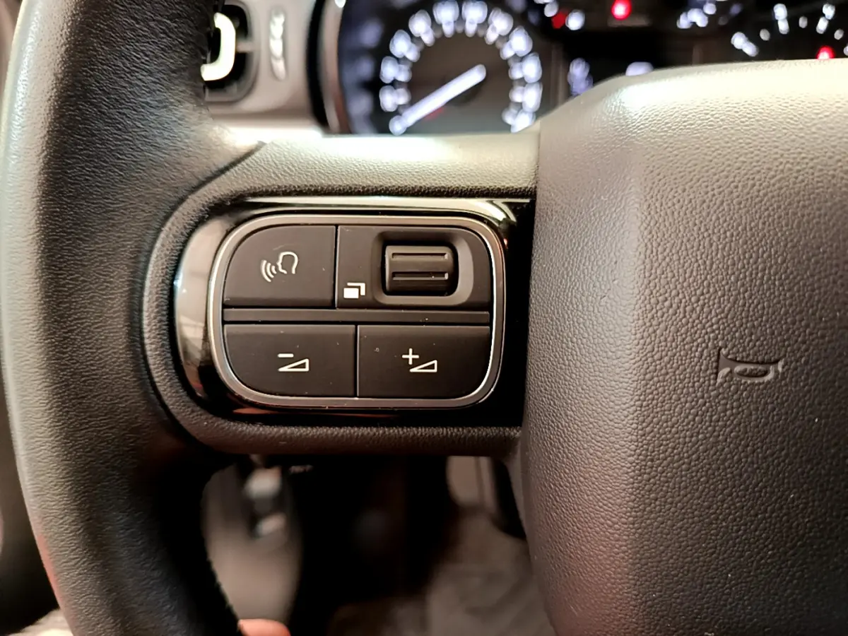 Gros plan sur les commandes gauche du volant cuir noir de la Citroën C3 Shine 2019, avec tableau de bord flou en arrière-plan.