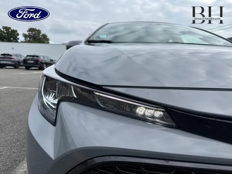 Gros plan sur le phare avant gauche d'une Toyota Corolla 122h Dynamic Business gris Manhattan métallisé sous ciel nuageux.
