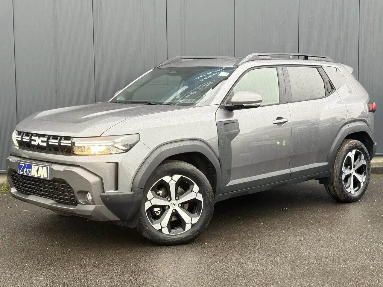 Dacia Duster gris schiste en 3/4 avant droit, avec jantes alliage diamantées et barres de toit noires.