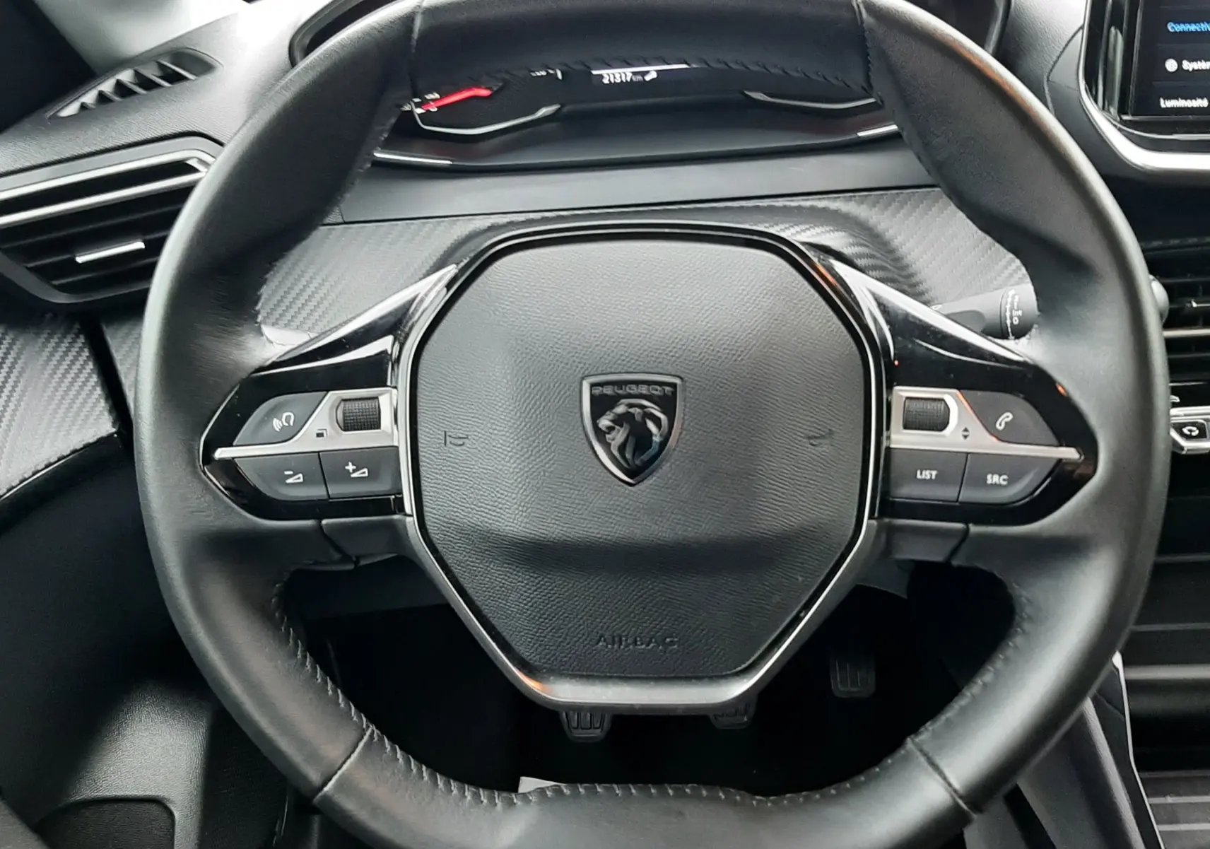 Vue rapprochée du volant noir de la Peugeot 208 2024, avec tableau de bord numérique et détails en carbone gris.