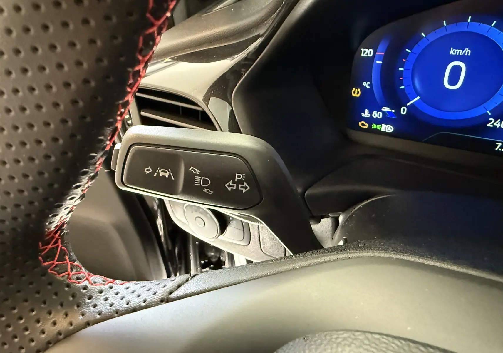 Gros plan sur la manette de clignotants à gauche du volant perforé noir avec surpiqûres rouges, tableau de bord numérique allumé.