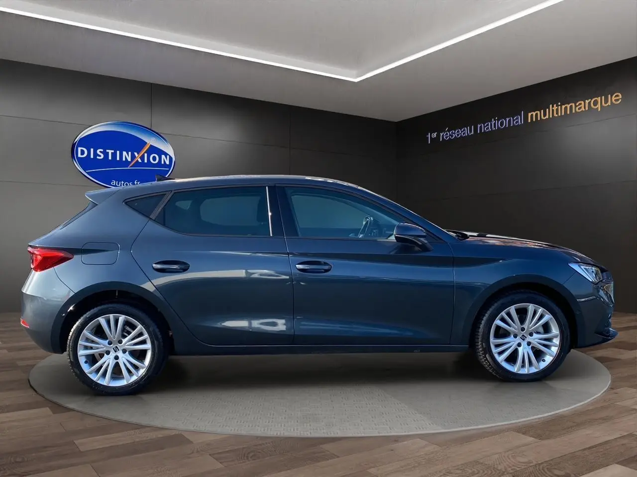 Profil droit d'une SEAT Leon IV gris magnetic avec jantes alu 17 pouces, dans un showroom moderne.