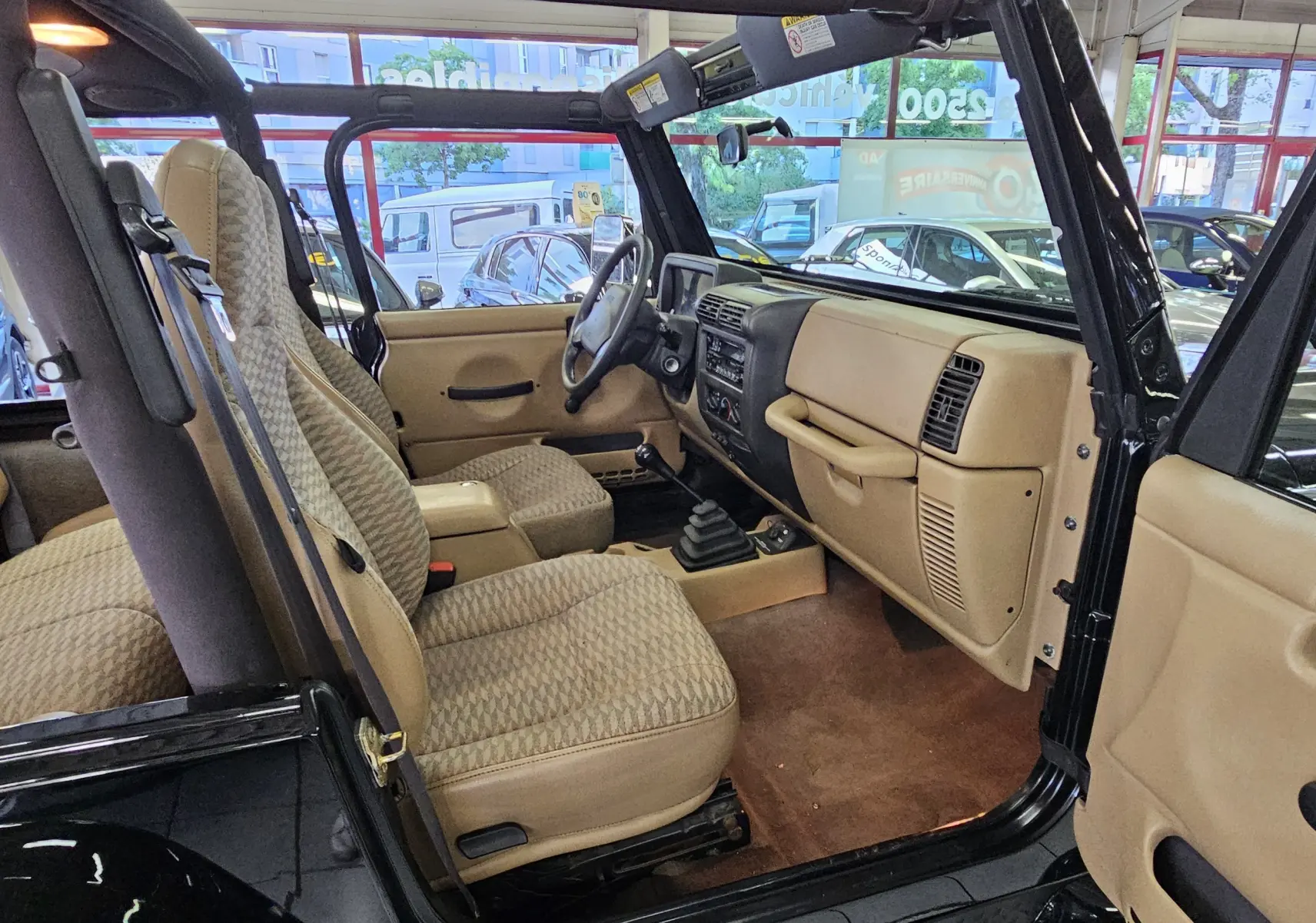 Intérieur beige vu côté conducteur d'une Jeep Wrangler noire de 2000 avec tableau de bord simple et boîte manuelle.