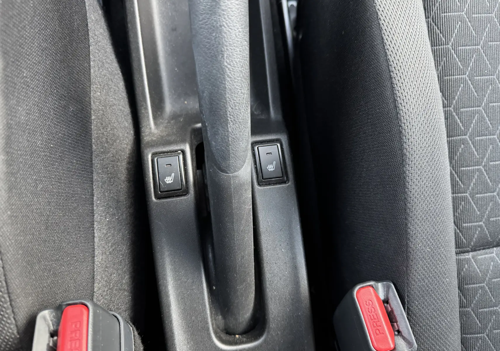 Gros plan sur la console centrale noire de la Suzuki Swift blanche 2025, montrant les boutons de sièges chauffants et les ceintures de sécurité.